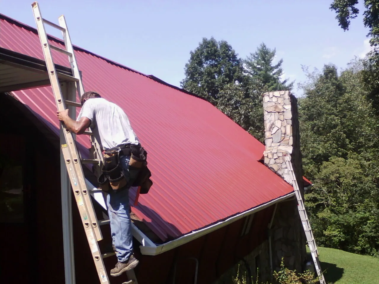 Roofer on a ladder beside a red metal roof and stone chimney — Soffit & Fascia Repair by Roofing Brothers in Port Murray
